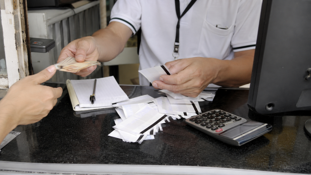  Operador realizando cobrança manual com dinheiro, tickets, calculadora e anotações sobre o balcão.
